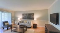 This single-floor space presents a thoughtfully designed living area that balances comfort with understated elegance. The room is finished with soft, neutral-toned walls in a muted sage-gray palette, creating a serene and welcoming ambiance. A continuous white chair rail runs along the perimeter, adding a classic architectural detail that grounds the space visually.

The living area is anchored by a light gray tufted loveseat, accessorized with an assortment of decorative pillows in complementary earth tones and soft blues. Above the sofa, two large ornate wall panels featuring intricate mandala-inspired metalwork serve as a striking focal point, lending a global, artisan aesthetic to the room.

Flanking the seating area are matching table lamps that cast a warm, inviting glow. A rustic wooden coffee table with a metal frame sits atop a neutral area rug, offering textural contrast. An ornate side table with a latticed design adds further character. Additional furnishings, including a wooden dresser and a decorative floral arrangement, complete the refined yet approachable aesthetic of this well-appointed space.