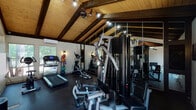 Located at 1117 Verde Dr in Colorado Springs, Colorado, this fitness center is a well-appointed single-floor facility designed to support a comprehensive workout experience. The space is immediately distinguished by its striking vaulted ceiling, finished in warm natural wood planking and accented by bold dark exposed beams that lend the room a rustic yet polished character. Track lighting runs along the ceiling, ensuring the entire space is brightly and evenly illuminated.

The fitness center is equipped with a full array of cardio machines, including a treadmill and elliptical trainer, as well as a multi-station cable strength machine and additional resistance equipment. Colorful medicine balls on a dedicated rack add a functional training element. Full-wall mirrors along one side of the room visually expand the space and allow users to monitor their form during workouts.

Rubber flooring throughout provides both durability and safety underfoot. A wall-mounted split air conditioning unit maintains a comfortable climate year-round. Natural light filters in through windows and large glass doors that open directly to an outdoor pool and courtyard area, creating an airy, connected atmosphere. A hygiene station is conveniently positioned near the entrance. The overall ambiance is clean, functional, and inviting, making this fitness center a practical and attractive amenity.