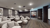 Located in Frisco, TX, this sophisticated private screening room delivers a true cinematic experience within a thoughtfully designed, single-floor space. The main theater area is anchored by a large projection screen mounted on a dark accent wall, paired with a ceiling-mounted projector that ensures a high-quality viewing experience. The seating arrangement features an array of plush, light gray swivel lounge chairs and soft sofas grouped around sleek round side tables with gold-accented legs, creating an intimate yet social atmosphere. The flooring throughout the screening area is a stylish patterned carpet with flowing gold and gray tones, adding warmth and visual depth to the dark, moody color palette of charcoal and slate gray walls. Abstract framed artwork adorns the walls, contributing to the space's contemporary aesthetic. Recessed lighting provides subtle, controllable illumination throughout. A dedicated concession area adjacent to the screening room elevates the experience further, equipped with a classic popcorn machine, a generous white quartz countertop, dark cabinetry, snack displays, and pendant lighting above — all set on striking marble-look tile flooring. Together, these spaces form a polished, amenity-rich entertainment venue ideal for private movie screenings and gatherings.