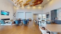 Located at 2620 Mall of Georgia Blvd in Buford, Georgia, this single-story community clubhouse delivers an impressive blend of modern design and welcoming functionality. The space is defined by its dramatic soaring ceilings accented with exposed wooden beams and striking starburst chandeliers, creating an airy, upscale atmosphere from the moment you enter.

The expansive main gathering area features wide-plank hardwood flooring throughout and is thoughtfully divided into distinct zones. A generous lounge area is furnished with plush blue velvet seating arranged around a linear fireplace set against a shiplap accent wall, with a mounted television above. Floor-to-ceiling multi-pane windows flood the interior with natural light and offer views of the outdoor surroundings.

An open dining and collaboration area provides large communal tables with contemporary seating, separated from adjacent spaces by a striking decorative laser-cut metal screen partition. The game room includes a shuffleboard table, warm caramel-toned seating, and a dramatic floor-to-ceiling stone-clad feature wall.

The kitchen area showcases bold navy-blue cabinetry, a sweeping marble backsplash, and a beverage station. Throughout, the design balances textural contrast — soft upholstery, natural wood, stone, and metal — creating a refined yet approachable environment ideal for community gatherings.