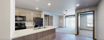 This single-floor apartment is located on Maeve Place in Waukee, Iowa, offering a practical and modern layout designed for comfortable everyday living. The open-concept kitchen serves as the heart of the unit, featuring light wood cabinetry, a center island with a double-basin sink and light countertops, and black appliances including a refrigerator, range, and over-the-range microwave. Recessed lighting illuminates the kitchen and adjoining areas, which transition seamlessly into the living space via light hardwood-style flooring. The living area is carpeted and features a ceiling fan, with access to what appears to be a patio or balcony offering natural light. Bedroom spaces are also carpeted and include windows with blinds that provide views of the surrounding neighborhood. Built-in shelving adds functional storage near interior doorways. The bathroom is well-appointed with a warm-toned wood vanity, light stone countertop, a wall-mounted mirror, a towel bar, and light wood-style flooring. A dedicated laundry area houses a full-size washer and dryer set beneath wire shelving that spans the width of the space, offering additional storage. The unit combines neutral tones and functional finishes throughout.
