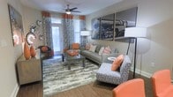 This single-floor space showcases a thoughtfully designed living area that balances bold style with functional comfort. The room is anchored by wood-look flooring and layered with a textured area rug, adding warmth and dimension underfoot. A contemporary gray sofa takes center stage, dressed with chevron and solid orange throw pillows that echo the room's vibrant color palette. Complementing accent chairs — one patterned in blue and white, another in solid orange — add visual interest and flexible seating options throughout the space.

A large-scale black-and-white photographic print of a locomotive serves as a dramatic focal point on the main wall, lending an industrial-chic character to the room. A ceiling fan with a built-in light fixture provides both airflow and ambient lighting, supplemented by coordinating floor lamps positioned throughout. Floor-to-ceiling patterned drapes in orange and teal frame the windows, allowing natural light to filter through while reinforcing the space's bold aesthetic. The overall ambiance is energetic yet cohesive, with a carefully curated mix of textures, patterns, and accent colors throughout.