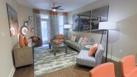This single-floor space showcases a thoughtfully designed living area that balances bold style with functional comfort. The room is anchored by wood-look flooring and layered with a textured area rug, adding warmth and dimension underfoot. A contemporary gray sofa takes center stage, dressed with chevron and solid orange throw pillows that echo the room's vibrant color palette. Complementing accent chairs — one patterned in blue and white, another in solid orange — add visual interest and flexible seating options throughout the space.

A large-scale black-and-white photographic print of a locomotive serves as a dramatic focal point on the main wall, lending an industrial-chic character to the room. A ceiling fan with a built-in light fixture provides both airflow and ambient lighting, supplemented by coordinating floor lamps positioned throughout. Floor-to-ceiling patterned drapes in orange and teal frame the windows, allowing natural light to filter through while reinforcing the space's bold aesthetic. The overall ambiance is energetic yet cohesive, with a carefully curated mix of textures, patterns, and accent colors throughout.