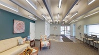 This well-appointed single-floor commercial space is designed to be both functional and inviting. The main entrance opens into a generously sized lobby featuring a striking multi-toned slate tile floor in warm earthy hues, transitioning seamlessly into carpeted areas. Exposed ceiling beams painted in slate gray complement the rows of modern pendant fluorescent fixtures and track lighting, creating a bright and well-lit environment throughout.

Adjacent to the entry is a comfortable lounge area, defined by a bold teal accent wall adorned with decorative artwork. This seating nook features a cream-toned sofa, patterned upholstered armchairs, a round wood coffee table, and a side table stocked with informational materials — ideal for welcoming visitors. Glass block windows allow soft, diffused natural light to filter into this cozy corner.

Further into the space, a large open hall provides additional versatility, featuring a long built-in counter with bar-height seating along a window wall, a wall-mounted flat-screen display, and a side door offering direct outdoor access. The layout also includes several private rooms or offices accessible via interior doors, as well as a small utility station with cabinetry and a countertop. The overall design balances a professional atmosphere with warm, approachable décor.