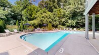 Located at 10 Highland Lake Cir in Decatur, Georgia, this single-level outdoor amenity space is centered around a generously sized freeform swimming pool with striking blue water, framed by an expansive concrete pool deck. The pool's organic, curved shape lends a resort-like quality to the setting, with multiple lounge chairs and patio seating arranged along its perimeter for relaxed outdoor enjoyment. Shaded dining areas with umbrellas provide relief from the sun, while a covered pergola structure with classic white columns anchors one side of the space, offering additional shelter and architectural interest. The surrounding landscape is thoughtfully designed, featuring a layered mix of evergreen shrubs, ornamental grasses, flowering plants, and mature deciduous trees that create a lush, green canopy overhead. Natural stone retaining walls and river rock border plantings add texture and depth to the garden beds. A wrought iron fence encloses the perimeter, providing privacy and security while maintaining an open, airy feel. The white-sided building exterior, complemented by stone accent walls, ties seamlessly into the well-maintained grounds, making this outdoor space a serene and inviting retreat in a verdant Decatur setting.