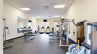 This fitness center offers a thoughtfully organized workout space designed to accommodate a wide range of exercise routines. The room features a drop-panel ceiling with recessed lighting that keeps the environment bright and functional, while large windows along one wall allow natural light to filter in and provide views of the surrounding greenery outdoors.

The cardio section is well-stocked with multiple treadmills, an elliptical trainer, and an upright stationary bike, all positioned near the windows to create an energizing workout environment. A wall-mounted flat-screen TV is conveniently placed above this area for entertainment during cardio sessions.

The strength training zone includes a multi-station cable machine and several Precor selectorized machines targeting key muscle groups. A full dumbbell rack with weights of varying sizes is neatly arranged in front of a large wall mirror, which amplifies the sense of space and allows for proper form monitoring. An adjustable weight bench completes the free weights area.

A dedicated functional training corner houses stability and medicine balls alongside additional cable equipment, offering versatility for core and balance workouts. The durable rubber flooring throughout provides safety and comfort underfoot. The overall aesthetic is clean and professional, with neutral cream-toned walls contributing to an open, welcoming atmosphere.