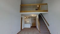 This two-story space is defined by its striking vertical volume, with dramatically tall ceilings that create an airy, open atmosphere throughout the main level. The ground floor features an open-concept layout where the living area flows naturally toward a compact kitchen tucked along the back wall. The kitchen is equipped with white cabinetry, an under-cabinet range hood, a cooktop, and a sink, offering a clean and functional workspace. Light-colored carpeting covers the main living area, contributing to a neutral, understated aesthetic throughout.

A carpeted staircase with a warm wooden banister leads to the upper loft level, which overlooks the main floor below through a full-length wooden balustrade with vertical spindles. This elevated second level adds functional square footage while reinforcing the sense of openness and height that characterizes the space. The entry door, finished in natural wood tones, anchors the main floor and provides a welcoming focal point. Track lighting on the main level and recessed fixtures on the upper level ensure the space is well-illuminated. Overall, the layout is practical and efficiently designed, making excellent use of vertical space.