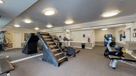 This fitness center offers a comprehensive and thoughtfully arranged workout environment across a single floor. The space is designed with functionality and comfort in mind, featuring neutral gray and beige walls accented by crisp white crown molding and chair-rail details that lend a polished, well-maintained appearance. Plush carpeted flooring extends throughout, providing a practical surface underfoot. Flush-mount circular ceiling lights ensure the space is consistently and evenly illuminated.

The equipment selection is extensive and varied. Cardio options include treadmills, elliptical trainers, a stationary bike, and a Jacob's Ladder climbing machine. Strength training is supported by a multi-function cable machine, a weight bench, and a rack of dumbbells. A Pepsi vending machine offers convenient refreshment access. Exercise accessories such as stability balls and resistance tools are also available.

Full-length mirrors along one wall visually expand the space and aid in form correction during workouts. Large windows and glass-paned doors allow natural light to filter in, with views suggesting a connection to an adjacent pool or courtyard area. A vaulted ceiling section near the entrance adds architectural interest. The overall ambiance is clean, functional, and welcoming, making this fitness center a well-rounded amenity for daily exercise routines.