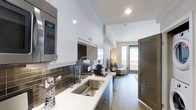 This single-floor space showcases a thoughtfully designed open-concept layout where the kitchen flows seamlessly into the living area. The kitchen is appointed with high-gloss white upper cabinetry that extends toward the ceiling, maximizing storage while creating a clean, modern aesthetic. A reflective dark tile backsplash adds contrast and visual depth, complementing the dark lower cabinetry and light countertops. A stainless steel double-basin sink paired with an industrial-style pull-down faucet serves as a functional focal point, while a built-in stainless steel microwave is neatly integrated into the cabinetry for a streamlined look. The galley-style kitchen leads the eye toward the living area, which benefits from generous natural light filtering through large windows fitted with horizontal blinds. The living space features hard flooring and is furnished with understated, contemporary pieces. A dark wood door with louvered panels adds a warm architectural detail. Recessed ceiling lighting throughout ensures the space remains bright and inviting, contributing to the overall modern, urban ambiance.