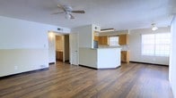 This single-floor residence offers a practical and well-organized layout designed for comfortable everyday living. The open main living area is anchored by rich, dark hardwood flooring that extends throughout, lending a warm and cohesive feel to the space. A ceiling fan with an integrated light fixture is centered overhead, providing both air circulation and ambient lighting beneath a textured popcorn ceiling.

The kitchen is defined by a wraparound peninsula with a white base and smooth countertop, creating a natural division between the cooking and living areas without closing off the floor plan. Warm-toned wood cabinetry lines the kitchen walls, offering ample storage, while a window above the sink allows natural light to filter into the space. A stainless steel refrigerator is visible at the far end of the kitchen, completing the functional setup.

A doorway visible from the main living area leads to an adjacent room with matching cabinetry and warm lighting, suggesting a utility or secondary prep space. HVAC vents are positioned thoughtfully throughout the ceiling and lower walls, ensuring consistent climate control. Dark baseboards provide a clean contrast against the light-colored walls, giving the interior a crisp, defined finish throughout. The overall atmosphere is open, functional, and move-in ready.