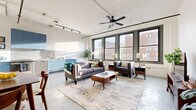 This single-floor urban loft showcases an open-concept layout with an industrial-modern aesthetic. Polished concrete floors run throughout the space, complementing the white-painted ceiling fitted with track lighting and a ceiling fan. Large, dark-framed windows dominate one wall of the living area, flooding the space with natural light and framing views of the surrounding cityscape.

The living area is generously proportioned, offering ample room for a full seating arrangement centered on a mid-century style coffee table. Adjacent to the living space, a dedicated area accommodates a media console and additional seating. The open-plan kitchen features a striking row of powder-blue cabinetry paired with light stone countertops, a stainless steel undermount sink with a brushed gold faucet, and a sleek backsplash—creating a vivid design focal point within the otherwise neutral palette.

A hallway connects the main living area to a bathroom with a walk-in shower and concrete floors, as well as a utility area housing a stacked washer and dryer. An electrical panel is conveniently accessible in the corridor. The overall ambiance is bright, airy, and distinctly urban, with a cohesive blend of industrial and contemporary design elements throughout.
