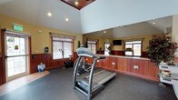The fitness center is a single-level space characterized by its striking vaulted ceiling with warm wood paneling at the peak, complemented by recessed lighting that creates a bright and energizing atmosphere. The elevated ceiling height adds a sense of openness, making the workout area feel spacious and airy. Walls are finished in a warm yellow tone with rich wood wainscoting that extends along the lower half, lending the space a polished, welcoming aesthetic. A large wall-length mirror runs along one side of the room, visually expanding the space and allowing users to monitor their form during exercise. The rubber flooring provides a durable, slip-resistant surface well-suited for fitness activities. The center is equipped with cardio machines including a treadmill and a stationary bike, with additional equipment visible in the mirror's reflection. Natural light filters in through multiple windows fitted with wood-toned blinds, enhancing the welcoming environment. A wall-mounted flat-screen television is also present, offering entertainment during workouts. Overall, the fitness center combines functional amenities with thoughtful design details for a comfortable exercise experience.