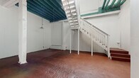 Located at 4401 San Leandro St in Oakland, CA, this two-story loft space blends industrial character with functional design across two well-proportioned floors. The ground floor greets visitors with richly textured polished concrete floors in warm terracotta tones, expansive white walls ideal for creative use, and a striking teal-painted exposed beam ceiling that adds a bold industrial accent. Pendant lighting hangs from the ceiling structure, and a white-railed staircase anchors the space, leading to the upper level. The ground floor also features direct exterior access through a solid door, offering flexibility for a variety of uses. Upstairs, the upper floor opens into a bright, airy space with light wood flooring, dramatic exposed wooden roof trusses, and a corrugated metal ceiling that reinforces the industrial aesthetic. Generous multi-pane windows flood the level with natural light. A well-appointed kitchen on this floor features white shaker cabinetry, dark countertops, full appliances including a gas range and refrigerator, and light wood flooring that carries throughout. An arched architectural detail adds a refined touch. The bathroom includes a full soaking tub and shower combo with clean white finishes. This distinctive space offers a compelling combination of raw industrial elements and modern livability.