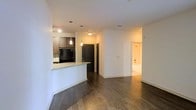 This single-floor residence showcases a thoughtfully designed open-concept layout that connects the kitchen, dining, and living areas into one cohesive space. The kitchen is outfitted with dark cabinetry, black appliances, and a granite-topped breakfast bar that serves as both a functional workspace and a casual dining area. Elegant pendant lighting hangs above the bar, adding warmth and a modern touch to the kitchen zone. Rich, dark hardwood-style flooring runs throughout the main living areas, creating a sense of continuity and sophistication. The crisp white walls provide a bright, neutral backdrop that enhances the overall sense of openness. A hallway visible from the main living area leads to the private quarters, including what appears to be a bathroom featuring a tub and shower combination. The layout is efficient yet comfortable, with clear sightlines from the kitchen through to the living space, making the interior feel expansive and well-connected. Recessed and flush-mount lighting throughout ensures the space remains well-lit. The overall ambiance is modern and clean, balancing contemporary finishes with a warm, inviting atmosphere suitable for everyday living.