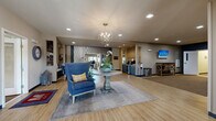This generously sized single-floor amenity center is thoughtfully designed to offer residents a variety of communal spaces, each with its own distinct character and function. The welcoming lobby serves as the heart of the facility, anchored by a statement round pedestal table topped with a lush floral arrangement and complemented by a striking orb chandelier overhead. Wood-look flooring, neutral tones, and blue accent seating establish a warm, contemporary aesthetic carried throughout the space.

Adjacent to the entry, a service counter area features built-in cabinetry, a beverage station, wall-mounted display screen, and rustic console furniture, providing a practical yet stylish transition point. The communal kitchen is well-appointed with granite countertops, a bold blue island with bar seating, white upper cabinetry, a mosaic tile backsplash, stainless appliances, and layered pendant lighting.

A comfortable lounge area showcases a linear gas fireplace framed by a stacked stone surround, a large wall-mounted television, deep navy accent walls, and generous seating arrangements on light hardwood flooring. A bright café-style dining room with high-top tables and large windows provides outdoor views and abundant natural light. The dedicated fitness center rounds out the facility, housing a comprehensive selection of cardio and strength equipment across patterned carpet flooring, with full-length mirrors enhancing the sense of space.