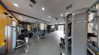 Located at 2158–2198 NW Fourth Ct in Boca Raton, FL, this single-floor fitness center is designed to support a full range of workout routines in a dynamic, energizing environment. The space is thoughtfully laid out to maximize functionality, with distinct zones dedicated to strength training, free weights, and cardiovascular exercise.

The strength training area is anchored by Hoist cable machines, a Smith machine, and adjustable benches, providing users with versatile options for resistance-based workouts. A full dumbbell rack lines the mirrored wall, with weights organized in ascending order for easy access. Floor-to-ceiling mirrors run along multiple walls, enhancing the sense of space and allowing users to monitor their form throughout their session.

The cardio zone is equipped with stair climbers, ellipticals, and treadmills, complemented by a wall-mounted television for entertainment. A garage-style glass door floods this section with natural light and provides views of the surrounding greenery, creating an open, airy atmosphere.

The interior design features bold orange and deep teal accent walls, large-scale fitness-themed wall murals, and durable rubber flooring throughout. Recessed lighting and ceiling fans ensure the space remains well-lit and comfortably ventilated. The overall ambiance is modern, motivating, and purpose-built for an active lifestyle.
