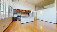 This urban loft suite at 675 North Main Street in Winston-Salem, NC showcases an industrial-modern aesthetic with soaring ceilings, exposed metal ductwork, and classic columns throughout the open living area. Rich bamboo hardwood floors flow from the spacious living area into the well-appointed kitchen, which features dark wood cabinetry, granite countertops, a center island, and a full suite of stainless steel appliances. Large industrial-style windows flood the space with natural light. The bedroom offers a comfortable retreat with a dedicated closet, while two full bathrooms include tile flooring and clean, modern fixtures. An in-unit laundry room with a stacked washer and dryer adds everyday convenience.