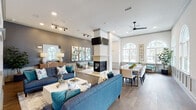 This well-appointed clubhouse is designed to serve as a premier communal gathering space, combining sophisticated design with practical amenities across a single, thoughtfully laid-out floor. The main lounge area is anchored by a striking double-sided fireplace, which serves as a central architectural feature dividing the space while maintaining an open, airy feel. Soaring ceilings with crown molding, recessed lighting, and a ceiling fan contribute to the bright and welcoming atmosphere. Large arched windows allow abundant natural light to fill the room, offering views of the surrounding community. The lounge is furnished with plush navy and teal sofas accented by decorative pillows, complemented by curated wall art and a warm wood-toned credenza with a statement mirror. An adjacent seating alcove with gold-framed accent chairs provides a more intimate setting beside the fireplace. A spacious dining area with a long communal table and upholstered seating flows seamlessly from the lounge. The fully equipped catering kitchen features dark cabinetry, granite countertops, stainless steel appliances including double wall ovens and a commercial-style range hood, and a generous curved island with bar stool seating beneath globe pendant lights. The consistent use of hardwood-style flooring, wainscoting, and a refined teal-and-gray color palette unifies the space with a cohesive, upscale aesthetic.