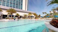 This resort-style pool complex offers a striking combination of modern architecture and tropical outdoor living. The centerpiece is a series of expansive, crystal-clear pools with vivid turquoise water, including a wide zero-entry leisure pool and a dedicated lap swimming lane. The pool deck is lined with tall, swaying palm trees and dotted with blue-umbrella shaded lounge seating, creating a vibrant, vacation-inspired atmosphere under open skies.

A decorative tiled water feature serves as a focal point within the leisure pool area, adding a Mediterranean-inspired architectural accent. The surrounding grounds are well-maintained, with lush tropical plantings and a smooth, light-toned hardscape that connects the various pool zones seamlessly.

The complex is anchored by a contemporary high-rise building whose glass facade towers above the pool deck, providing a dramatic architectural backdrop. A lower-level covered pavilion runs along one side, offering shaded access points directly to the pool area.

The interior entry space features polished marble-style tile flooring with inlaid border detailing, neutral walls, and recessed ceilings — conveying a clean, upscale finish. Glass double doors open directly to the pool grounds, framing views of palm trees and blue umbrellas, blurring the boundary between interior and outdoor resort spaces.