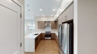 This single-floor space showcases a contemporary design aesthetic with a cohesive neutral color palette of warm beige tones, light wood-look flooring, and clean white trim throughout. The kitchen serves as a functional centerpiece, featuring wood-grain flat-front cabinetry extending to the ceiling for maximized storage, light quartz countertops, a stainless steel gas range with a wall-mounted hood, a built-in microwave, a full-size stainless steel refrigerator, and a peninsula with an undermount sink — all illuminated by recessed lighting and a pendant fixture. A large window brings in natural light and views of the surrounding area. The open living area benefits from a bank of oversized windows offering outdoor views, creating a bright and airy atmosphere with generous floor space. The hallways are well-organized, featuring white shaker-style double doors for closet storage and a recessed utility panel. Bedrooms are spacious with tall ceilings and dedicated ventilation. The bathrooms are refined, with dual-sink vanities topped in white quartz, warm wood cabinetry, large mirrors, and glass-enclosed showers finished in neutral tile. The overall design is clean, modern, and move-in ready.
