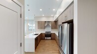 This single-floor space showcases a contemporary design aesthetic with a cohesive neutral color palette of warm beige tones, light wood-look flooring, and clean white trim throughout. The kitchen serves as a functional centerpiece, featuring wood-grain flat-front cabinetry extending to the ceiling for maximized storage, light quartz countertops, a stainless steel gas range with a wall-mounted hood, a built-in microwave, a full-size stainless steel refrigerator, and a peninsula with an undermount sink — all illuminated by recessed lighting and a pendant fixture. A large window brings in natural light and views of the surrounding area. The open living area benefits from a bank of oversized windows offering outdoor views, creating a bright and airy atmosphere with generous floor space. The hallways are well-organized, featuring white shaker-style double doors for closet storage and a recessed utility panel. Bedrooms are spacious with tall ceilings and dedicated ventilation. The bathrooms are refined, with dual-sink vanities topped in white quartz, warm wood cabinetry, large mirrors, and glass-enclosed showers finished in neutral tile. The overall design is clean, modern, and move-in ready.