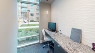 This single-floor professional space showcases a refined, contemporary design throughout. The entry corridor features light-toned tile flooring, crisp white baseboards, and floor-to-ceiling glass walls that invite natural light and offer views of the surrounding outdoor greenery and streetscape. The clean, neutral palette of soft gray walls creates a calm, polished atmosphere from the moment of arrival.

The central lounge or waiting area serves as a focal point of the space, anchored by a warm wood accent wall that provides a rich textural contrast to the otherwise modern finishes. A large, bold abstract artwork in deep blue and white commands attention above a plush gray sectional sofa accented with purple throw pillows. Sleek side tables with metal bases and wood tops complete the seating arrangement. A wall-mounted display screen adds a functional, connected element to the area.

A dedicated workstation nook offers a built-in granite-topped desk, a task chair, and a desktop computer, positioned alongside expansive floor-to-ceiling windows that frame outdoor views. A textured wave-patterned accent wall adds visual interest nearby. Glass-walled partitions throughout maintain an open, airy feel while defining distinct functional zones within the space.