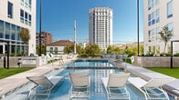 This expansive outdoor amenity space is designed to deliver a resort-like experience within an urban setting. At its centerpiece sits a generously sized swimming pool with crystal-clear water, flanked by wide concrete pool decks outfitted with rows of sleek white lounge chairs—ideal for sunbathing and relaxation. A separate spa offers a dedicated hydrotherapy area, complete with accessible entry equipment and glass panel railings that frame sweeping views of the surrounding cityscape and distant mountain range.

Patches of lush artificial turf provide a soft, green contrast to the light-toned hardscape, while a shaded pergola structure offers a comfortable outdoor seating area for gatherings or casual lounging. Raised planters with manicured greenery and young trees add natural texture throughout the space, contributing to a well-considered landscape design.

The deck is enclosed by the building's modern multi-story facade, characterized by large-format windows, private balconies, and a clean, contemporary aesthetic using neutral tones with dark accent panels. The open-air layout allows for abundant natural light throughout the day. Together, these elements create a polished and functional outdoor retreat that balances comfort, design, and urban convenience.