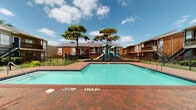 This outdoor amenity area serves as a central gathering space within a brick-constructed residential community. The centerpiece is a generously sized swimming pool with clear turquoise water, framed by a classic red brick paver deck that provides ample space for lounging. Pool depth markers are clearly posted, and entry is facilitated by stainless steel handrails. A dedicated wading pool sits adjacent, offering a safer option for younger children. Surrounding the pool area is a black iron perimeter fence, ensuring safety while maintaining open sightlines to the landscaped grounds beyond.

A colorful multi-element playground structure, complete with slides and climbing features, is positioned nearby, making this area family-friendly. A pergola-covered structure along one side of the deck provides shaded seating and houses posted pool rules and safety signage. Mature trees, including distinctive ornamental pines, are scattered throughout the community, providing natural shade and contributing to a pleasant, park-like atmosphere.

The residential buildings surrounding the amenity area are two-story brick structures featuring balconies, exterior staircases, and landscaped garden beds. The combination of open sky, mature greenery, and well-maintained hardscaping creates an inviting and functional outdoor environment suited for both recreation and relaxation.