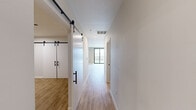 This well-appointed 1-bedroom, 2-bathroom single-floor residence offers a thoughtfully designed open layout with a clean, contemporary aesthetic. Light hardwood-style flooring runs throughout, complementing the crisp white walls and recessed lighting that give the space a bright, airy feel.

The combined kitchen and living area serves as the heart of the unit. The kitchen is outfitted with stainless steel appliances — including a refrigerator, electric range, built-in microwave, and dishwasher — paired with white shaker-style cabinetry, a white subway tile backsplash, and light quartz countertops. A matte black faucet with an undermount sink adds a modern contrast to the clean palette.

The spacious living area benefits from a large floor-to-ceiling window with access to a private balcony, flooding the room with natural light and offering outdoor views. The hallways are punctuated by a striking set of double sliding barn doors with black hardware, adding a stylish industrial-modern touch.

The primary bedroom features a clothing rod for storage and continues the consistent hardwood flooring. The two bathrooms are finished with clean, neutral tones — one includes a walk-in shower with chrome fixtures, while both feature wood-toned vanities topped with white quartz and black-framed mirrors. A utility room completes the functional layout.