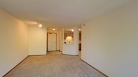 This single-floor residence offers a clean, functional layout designed to maximize open living space. The main living area features neutral wall-to-wall carpeting and light-toned walls that create a bright, airy atmosphere throughout. A centrally positioned half-wall with a rounded base separates the living area from the kitchen, providing an open pass-through that enhances connectivity between spaces while maintaining definition. The kitchen is equipped with warm wood-toned cabinetry on both the upper and lower levels, a sink positioned beneath a window, and tile or hard surface flooring that contrasts neatly with the carpeted living space. The entryway, accessible through a solid paneled door, is well-lit by a ceiling-mounted flush light fixture and accented by adjustable track lighting overhead, ensuring the space feels welcoming upon entry. Baseboard trim in a complementary wood finish runs throughout, adding a cohesive finishing touch to the interior. The open floor plan, combined with consistent neutral tones and practical finishes, gives the space a versatile and livable character suited to a variety of furnishing styles.