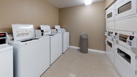 Located at 260 Marcella Rd in Hampton, Virginia, this single-floor community amenity building at The Township offers residents convenient access to essential shared facilities. The space is finished with neutral, warm-toned walls and durable vinyl tile flooring throughout, creating a clean and functional environment.

The fitness center is a standout feature, visible through large glass windows and a glass-paneled door from the main corridor. It is equipped with a variety of cardio machines, including treadmills, ellipticals, and a stationary bike, all arranged on a dark rubberized floor. Natural light filters in through exterior-facing windows, lending an open and energizing atmosphere to the workout area.

The laundry room provides a practical, well-organized space lined with multiple top-loading washing machines and additional dryers, offering ample capacity for residents. The room is brightly lit, with textured ceilings and easy-to-maintain flooring. A folding counter and waste bin add to the room's functionality.

Connecting these spaces is a wide, well-lit corridor featuring direct exterior door access, emergency safety equipment, and a dark granite-topped counter surface for added convenience. Signage throughout reflects the managed, community-oriented nature of the facility. The overall design prioritizes practicality and ease of use for all residents.