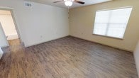This single-story space presents a clean, straightforward layout designed for practical living. The main room is an open, generously sized area finished with durable wood-look plank flooring that runs throughout, offering a warm, natural aesthetic while remaining easy to maintain. The flooring features a realistic wood grain pattern in earthy tones, complementing the neutral cream-colored walls that keep the space feeling bright and airy.

A ceiling fan with an integrated light fixture is centrally mounted, providing both air circulation and ambient lighting. A window fitted with horizontal blinds allows natural light to filter into the room while offering privacy control. Multiple electrical outlets are positioned along the walls, ensuring functional flexibility for furniture arrangement and everyday use.

The white baseboards provide a clean contrast against the flooring and walls, contributing to a tidy, well-finished appearance. The overall ambiance of the space is simple and neutral, making it highly adaptable to a variety of uses and décor styles. The absence of textured or decorative elements keeps the focus on the clean lines and practical features of the interior.