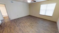 This single-story space presents a clean, straightforward layout designed for practical living. The main room is an open, generously sized area finished with durable wood-look plank flooring that runs throughout, offering a warm, natural aesthetic while remaining easy to maintain. The flooring features a realistic wood grain pattern in earthy tones, complementing the neutral cream-colored walls that keep the space feeling bright and airy.

A ceiling fan with an integrated light fixture is centrally mounted, providing both air circulation and ambient lighting. A window fitted with horizontal blinds allows natural light to filter into the room while offering privacy control. Multiple electrical outlets are positioned along the walls, ensuring functional flexibility for furniture arrangement and everyday use.

The white baseboards provide a clean contrast against the flooring and walls, contributing to a tidy, well-finished appearance. The overall ambiance of the space is simple and neutral, making it highly adaptable to a variety of uses and décor styles. The absence of textured or decorative elements keeps the focus on the clean lines and practical features of the interior.
