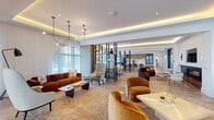 This elegantly designed single-floor space blends refined materials with contemporary interiors to create a cohesive and luxurious environment. The entry corridor immediately establishes a striking tone, lined with dramatic veined marble cladding and herringbone hardwood flooring beneath a series of sculptural pendant lights. The open-plan lounge area is expansive and bathed in natural light, courtesy of floor-to-ceiling windows dressed in sheer white drapery. Curved, jewel-toned furnishings in rust, amber, and gold add warmth against the neutral palette, while layered ambient lighting — including cove-lit tray ceilings and artistic chandeliers — creates an inviting atmosphere. A sleek open kitchen anchors one end of the space, showcasing a generous waterfall-edge marble island, light wood cabinetry, matte black fixtures, and warm recessed lighting overhead. Adjacent to the lounge, a formal dining area seats a large group around an oval wood table beneath a globe-pendant chandelier, framed by sheer-curtained windows. A dedicated game room features a full-size billiards table, built-in cabinetry, a wall-mounted display, and a coffered ceiling with ambient uplighting. Throughout, herringbone hardwood floors, curated artwork, and bold marble accents unify the space with a polished, design-forward aesthetic.