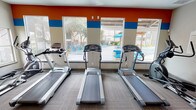 This fitness center is a thoughtfully designed, single-floor workout facility that offers a comprehensive range of exercise equipment to support diverse fitness routines. The interior is distinguished by bold orange and blue horizontal accent bands along the upper walls, providing an energetic and motivating atmosphere against a neutral base palette.

The cardio section is anchored by a row of treadmills and elliptical trainers positioned along large windows that face the outdoor pool area, allowing natural light to fill the space and providing pleasant views while exercising. Additional cardio equipment includes upright and recumbent stationary bikes, catering to a variety of fitness preferences.

The strength training area features a multi-station cable and weight machine, along with a leg press/extension unit, offering users options for targeted resistance training. A dedicated free weights section includes a full dumbbell rack with a wide weight range, positioned in front of a large wall mirror that enhances the sense of space and allows users to monitor their form.

The flooring throughout consists of durable patterned carpet tiles, while recessed lighting and ceiling fans ensure a comfortable environment. A glass-paneled entry door with safety equipment nearby provides easy, secure access. Overall, the fitness center delivers a clean, functional, and well-lit space suited for full-body workouts.