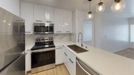 Located at 10343 San Diego Mission Rd in San Diego's Mission Valley neighborhood, this single-floor unit showcases a clean, modern aesthetic throughout. The open-concept layout connects the living area to the kitchen, which serves as the design centerpiece. The kitchen features glossy white cabinetry with matte black hardware, a striking marble-style tile backsplash, quartz countertops, and a full suite of stainless steel appliances including a range, over-the-range microwave, refrigerator, and dishwasher. Globe pendant lights add a contemporary finishing touch above the kitchen peninsula, which also functions as a breakfast bar. The living area is finished with light wood-look flooring and is accented by a ceiling fan. Carpeted bedrooms offer a comfortable retreat, each with windows dressed in blinds that allow natural light to filter in, with views of the surrounding neighborhood. The primary bedroom includes mirrored closet doors and connects to a bathroom featuring quartz countertops, a white vanity, and well-placed mirrors. Additional storage closets are visible throughout the layout. The overall design palette of crisp whites, neutral tones, and modern fixtures creates a bright, cohesive, and move-in-ready living environment.