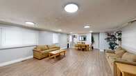Located at 5220 Luzzane Dr in Indianapolis, IN, this single-floor community space offers a well-organized layout designed for both relaxation and activity. The main gathering area is expansive and open, featuring durable wood-look flooring, neutral gray walls, and multiple flush-mount ceiling light fixtures that provide consistent, even illumination throughout. Large windows fitted with blinds allow natural light to filter into the space while maintaining privacy.

The lounge area is furnished with comfortable seating, coffee tables, and side tables, while decorative potted plants and framed artwork add warmth to the environment. Adjacent to the lounge, a dining area accommodates a full table and chair set, making it well-suited for communal meals or group meetings. A mirror and framed art on the walls enhance the sense of openness in this section.

A compact kitchenette is equipped with white cabinetry, a stainless steel sink, countertop space, a microwave, and a dishwasher — providing convenient food preparation and clean-up capabilities. The exercise room is outfitted with multiple cardio machines, including treadmills and other fitness equipment, offering a dedicated space for physical wellness. Throughout the space, the clean, functional design and cohesive aesthetic make it a practical and welcoming environment for community use.