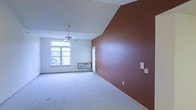 This single-floor space makes a strong visual impression through its thoughtful design details and open layout. The main room features a vaulted ceiling that adds a sense of height and airiness, complemented by a ceiling fan that provides both functional ventilation and a clean aesthetic touch. A striking terracotta-toned accent wall anchors the room, creating a warm focal point that contrasts with the surrounding white walls and light-colored carpet underfoot. Natural light filters in through a large multi-pane window, offering views of surrounding greenery and brightening the interior throughout the day. A wall-mounted HVAC unit ensures year-round climate comfort. The doorway visible along the accent wall suggests additional adjoining space, adding to the overall functionality of the layout. The combination of neutral flooring, clean white ceilings, and the bold accent wall gives the space a balanced, inviting atmosphere that is both practical and visually engaging. Overall, this space offers a straightforward yet characterful environment, well-suited for a variety of uses.