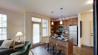 This single-floor space showcases an open-concept layout that seamlessly connects the kitchen and living area, creating a functional and inviting environment. The kitchen is a standout feature, equipped with stainless steel appliances including a refrigerator, microwave, and range. Warm-toned wood cabinetry provides ample storage, complemented by a tasteful tile backsplash that adds texture and visual interest. The kitchen island, topped with dark granite-style countertops, doubles as a casual dining space with industrial-style bar stools, making it ideal for both meal preparation and entertaining. Pendant lighting hangs above the island, while a track lighting system illuminates the kitchen workspace effectively. Hardwood-style flooring runs throughout, adding warmth and continuity to the space. Natural light flows generously through large windows fitted with horizontal blinds, brightening the living area where a plush area rug and accent chair contribute to a comfortable atmosphere. The presence of a glass-panel door provides access to the outdoors while further enhancing the natural light within the space.