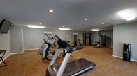 This fitness center in Baton Rouge, Louisiana offers a spacious, single-floor layout designed to accommodate a well-rounded workout experience. The room features neutral gray walls, patterned carpet tile flooring, and a combination of recessed can lighting and flush-mounted fluorescent fixtures that illuminate the space evenly throughout.

The cardio section is equipped with multiple treadmills, elliptical trainers, and a stationary bike, each positioned with ample space between units for comfortable use. Wall-mounted flat-screen televisions are installed at strategic viewing angles, allowing users to stay entertained during cardio sessions. The strength training area includes a cable crossover machine, a multi-function cable station, a lat pulldown machine, a preacher curl bench, and a dedicated free weight bench, providing a comprehensive range of resistance training options.

Climate control is maintained through multiple wall-mounted mini-split air conditioning units positioned around the perimeter of the room, an important feature given Baton Rouge's warm climate. Two sets of glass panel exterior doors line one wall, allowing natural light to filter into the space and providing direct access to the outdoors. The overall ambiance is clean, functional, and well-maintained, making it a practical facility for daily fitness routines.