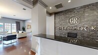 The Glenbrook clubhouse is a expansive single-floor community facility thoughtfully designed to serve as a hub for residents. The entrance greets visitors with an elegant reception desk finished in white wainscoting and a dark granite countertop, set against a striking stacked-stone accent wall bearing the Glenbrook logo. Polished concrete floors in a rich amber tone flow seamlessly throughout the common areas, reflecting natural and recessed lighting for a warm, upscale ambiance.

The open living and dining area centers around a bold white fireplace with mounted television, complemented by comfortable lounge seating and direct access to the outdoors through glass double doors. Adjacent to this space, a large kitchen and bar area features a wraparound counter with a dark granite surface and modern bar stools, ideal for social gatherings.

The business center offers a productive workspace with multiple computer workstations and stylish task seating, alongside a dedicated private conference room equipped with a wall-mounted display and natural window light. A well-appointed fitness center rounds out the amenities, housing a comprehensive selection of strength and cardio equipment set against bold blue accent walls. A laundry room and connecting hallway complete this thoughtfully planned community space.