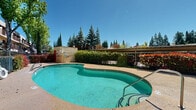 Located at 233 Clark Ave in Yuba City, California, this outdoor communal space centers around a freeform swimming pool that serves as the focal point of the property. The pool's kidney-shaped design features striking teal water and is framed by a generous concrete sundeck ideal for lounging under the California sun. Surrounding the pool area is mature, well-maintained landscaping, including tall evergreen trees, vibrant red-leafed shrubs, and seasonal flowering plants that provide both privacy and a lush, park-like atmosphere. A covered pergola structure along one edge of the pool deck offers shaded seating and an area for outdoor relaxation. The pool area is enclosed by decorative iron fencing, enhancing both security and aesthetic appeal. Overlooking the entire outdoor space is a multi-story residential building featuring individual private balconies, bold blue entry doors, and a neutral stucco exterior accented with awnings. The balconies are accessible via exterior staircases, and many are adorned with potted plants, adding character and warmth. Tall birch and willow trees punctuate the courtyard, contributing to the serene, shaded environment. The space benefits from abundant natural light and an open-sky setting, making it an inviting and vibrant outdoor retreat for residents.