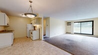 This single-floor unit offers a straightforward, functional layout with a clear distinction between the kitchen and living areas. The kitchen is equipped with white cabinetry, a coil-top electric range, a full-size refrigerator, and a under-cabinet range hood. Tile flooring runs through the kitchen, transitioning to wall-to-wall carpet in the open living area. A pendant light fixture illuminates the kitchen-living transition zone, while a wall-mounted air conditioning unit and a sliding glass door with outdoor views define the far end of the living space, allowing natural light to fill the room. Baseboard heating units run along the perimeter of multiple rooms, providing consistent warmth throughout. The bedrooms are carpeted and feature neutral yellow-toned walls, large windows with outdoor views of greenery, and built-in closets with paneled bifold doors. The bathroom includes a full-size soaking tub with a shower combo set within a clean, white surround with chrome fixtures. Overall, the space maintains a simple, neutral aesthetic with practical finishes and a functional floor plan suited for everyday living.