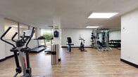 This fitness center is a thoughtfully designed exercise facility spread across a single floor, offering a comprehensive range of workout options in a clean and functional environment. The space features light wood-style flooring throughout, white walls, and recessed skylight-style panel lighting that creates a bright, energizing atmosphere.

The main workout area is generously proportioned, accommodating a diverse selection of equipment without feeling crowded. Cardio options include multiple treadmills and a stationary spin bike, with the treadmill area benefiting from large floor-to-ceiling windows that provide natural light and views of a landscaped outdoor greenspace. An elliptical trainer is also available for low-impact cardio workouts.

The strength training section is well-appointed, featuring a multi-function cable and weight machine, a dedicated free weights area with a dumbbell rack and exercise mat, and a weight bench. A wall-mounted mirror in the strength zone allows users to monitor their form during exercises.

An additional cable machine with attachments offers further versatility for resistance training. The facility also includes a compact, private restroom with a modern round mirror, a wall-mounted sink, and a toilet, finished in the same clean, minimalist aesthetic as the rest of the space. Overall, the fitness center provides a practical and well-maintained environment suitable for a full-body workout routine.