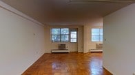 This single-floor unit is characterized by its warm, classic parquet hardwood flooring, which adds timeless charm to the open living area. The space is well-lit by multiple windows, including a pair of larger windows at the front that frame views of mature trees outside, inviting abundant natural light into the interior. A door situated between the front windows provides direct access to the outdoors. An additional window on the adjacent wall further brightens the space and enhances ventilation. The walls are painted in a clean, neutral white, contributing to an open and airy feel. Practical amenities include wall-mounted air conditioning units and baseboard heating radiators positioned beneath the windows, ensuring year-round comfort. The layout is straightforward and open, offering flexible possibilities for furniture arrangement. The combination of the rich parquet floors, neutral walls, and generous natural light creates a functional yet inviting atmosphere suited to a variety of uses.