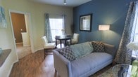 This single-floor residence seamlessly integrates a living area and dining space into one cohesive, open layout. The living area is anchored by a spacious grey sofa accented with decorative throw pillows in coordinating teal and white patterns, complemented by a plush area rug beneath. A bold teal accent wall adds depth and visual interest to the space, contrasting elegantly against the neutral cream-toned surrounding walls. A framed wall mirror enhances the sense of openness, while a tall floor lamp provides warm ambient lighting.

The dining area sits adjacent to the living space, featuring a compact round dark-wood table paired with two white upholstered chairs, ideal for intimate dining. A window dressed with patterned curtains allows natural light to filter into the room, brightening the space and highlighting the rich hardwood flooring that runs throughout. Decorative branches in a tall vase add a natural, artistic touch near the dining area.

Overall, the space presents a well-curated, contemporary aesthetic, balancing functionality with thoughtful design details, creating a welcoming and comfortable environment.