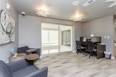 This single-floor space is designed as a functional and stylish business lounge, seamlessly combining collaborative and individual work areas within a contemporary setting. The layout flows across several distinct zones, each thoughtfully appointed to serve different professional needs.

The conference room is a standout feature, anchored by a warm-toned rectangular wooden table surrounded by quilted, upholstered chairs with sleek metal frames. A sculptural pendant light in amber tones hangs above the table, adding an artistic focal point. Floor-to-ceiling windows along one wall flood the room with natural light and provide outdoor views of surrounding greenery, creating an inviting atmosphere for meetings.

Adjacent to the conference area, a compact workstation nook houses two desktop computer setups along a dark wood desk, ideal for focused individual tasks. Glass partition walls contribute to an open, transparent feel while maintaining functional separation between areas.

The central lounge area features two cushioned armchairs paired with a small round side table, accented by abstract wall art and sculptural ceiling light fixtures. Neutral-toned carpet tiles throughout the space add texture and cohesion. Large windows continue to allow natural light to permeate the lounge, reinforcing the space's bright, professional, and welcoming ambiance.