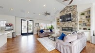 Located at 125 N Mill Rd in Vineyard, Utah, this single-floor communal space combines comfort and functionality across an open, welcoming layout. The living room serves as the centerpiece, showcasing a dramatic floor-to-ceiling natural stone fireplace flanked by built-in shelving with cabinetry below. Vaulted ceilings with receding angles amplify the sense of openness, while ceiling fans and recessed lighting add to the airy atmosphere. Large French doors and windows flood the space with natural light and offer views of the surrounding exterior. Warm hardwood-style flooring runs throughout, unifying each area seamlessly. The kitchen is thoughtfully designed with white shaker cabinetry, granite countertops, a mosaic tile backsplash, stainless steel appliances, and a central island with bar seating—ideal for casual gatherings. A beverage station adds a convenient communal touch. The adjacent dining area features granite-topped bar tables with stools positioned near expansive windows, creating a bright and social setting. Coastal-inspired wall art and lush potted plants contribute to the inviting, relaxed ambiance of this well-designed community gathering space.