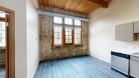 Located at 550 W Western Ave in Muskegon, Michigan, this 1-bedroom, 1-bathroom unit showcases a compelling blend of historic industrial character and contemporary updates. Exposed brick walls, warm wooden ceiling beams, and visible ductwork give the space a distinctive loft aesthetic throughout.

The living area features smooth, light-toned luxury vinyl plank flooring and benefits from an abundance of natural light streaming through large, wood-framed multi-pane windows. The high ceilings amplify the sense of openness, making the space feel larger than its footprint suggests.

The kitchen is outfitted with modern gray cabinetry, a full suite of stainless steel appliances including a refrigerator, range, microwave, and dishwasher, and clean countertops — all set beneath soaring ceilings with industrial-style overhead lighting.

The generously sized bedroom continues the exposed brick and beam aesthetic, with soft carpet flooring and oversized windows that frame views of the surrounding neighborhood. A dedicated closet provides practical storage, maintaining the clean lines of the bedroom.

The bathroom is finished in a clean, neutral palette, featuring wood-look vinyl flooring, a full tub-shower combination, and light wood-toned cabinetry. The overall ambiance of this unit is one of urban character rooted in the building's history, thoughtfully refreshed with modern conveniences.