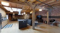 This single-floor industrial loft showcases a masterful blend of historic character and contemporary design. The open-concept layout is defined by soaring exposed timber beam ceilings, which run the length of the space and create a warm, structural aesthetic overhead. Visible ductwork and track lighting complement the industrial framework while providing functional illumination throughout.

The expansive main living area is anchored by a large dark stone kitchen island with bar seating, positioned beneath pendant lights that draw the eye upward to the dramatic ceiling. The kitchen flows seamlessly into the living area, which features a generous sectional sofa and a wall-mounted flat-screen television set against a bold exposed brick wall — a defining feature that adds texture and warmth throughout the space.

Heavy timber post-and-beam columns punctuate the interior, reinforcing the building's historic bones while defining zones within the open floor plan. Light hardwood flooring runs throughout, offering a bright contrast to the richness of the brick and wood above. Natural light enters through skylights set into the ceiling structure, further enhancing the airy yet grounded atmosphere. The overall ambiance is one of refined industrial character — raw, spacious, and thoughtfully designed.